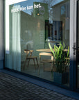 A glass-fronted room with two wooden chairs, an Ethnicraft Torsion Dining Table - Oak highlighting fine woodworking, a radiator, potted snake plant, and white curtains. Reflections and text on the window read: “HOEK. Hier kan het.”.