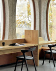 A modern study area features wooden desks and Ethnicraft Bok Dining Chairs in Black Oak with Leather, set by large arched windows filled with autumn light. Open notebooks and a cup rest on the solid oak desk.