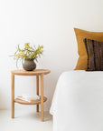 A minimalist bedroom features the Ethnicraft Bok Side Table - Oak, adorned with a vase of yellow flowers and books beneath. Beside it sits a bed dressed in white linens with mustard and brown pillows against a white wall.
