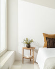 A minimalist bedroom showcases the Ethnicraft Bok Side Table - Oak, adorned with a yellow flower vase and books, positioned next to a bed with white bedding and amber-brown pillows as sunlight pours in through a large window.