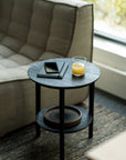 A compact, round Bok Side Table - Black by Ethnicraft sits next to a beige sofa, holding a tablet, smartphone, and orange juice; its lower shelf displays stacked plates as sunlight streams through the nearby window.