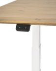 A close-up of the Ethnicraft Bok Adjustable Desk - Oak shows its white legs and a small black control panel with up/down arrows beneath the tabletop for height adjustment.
