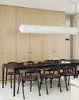A modern dining room with an Ethnicraft Bok Extendable Dining Table in brown oak, six matching chairs, a light rug, minimalist beige built-in cabinets, and a long white pendant light hanging above the tableware.