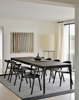 A modern dining room with a black rectangular table, six black chairs, an Ethnicraft Dots Kilim Rug (100% wool), minimalist vases, abstract wall art, a tall lamp, and a large window with white curtains letting in natural light.