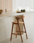 The Bok Counter Stool by Ethnicraft, with its modern solid oak frame and curved backrest, sits at a light kitchen island surrounded by minimalist light wood cabinets, artichokes, a glass of water, and a bowl on the counter.
