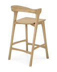 The Bok Counter Stool by Ethnicraft is a modern solid oak piece with a curved backrest, angled legs, and a natural light wood finish. Its contoured seat and minimalist design reflect Ethnicraft's signature style.