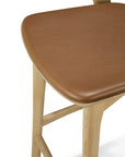 A close-up of the Ethnicraft Bok Barstool - Oak, featuring a light brown faux leather cushioned seat and solid oak legs, shown against a white background.