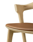 Close-up of the Ethnicraft Bok Barstool in solid oak, featuring a curved backrest and a brown leather seat cushion, shown against a white background.