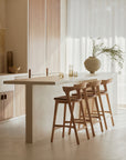 A minimalist kitchen features a white island, three Ethnicraft Bok Barstools in solid oak, sheer curtains, wooden cabinets, and a vase with green branches on the counter, all bathed in soft natural light.