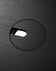 A close-up of the circular cable management hole, partially covered, on the Ethnicraft Bok Adjustable Desk in Black Oak, with a white background visible through the opening.