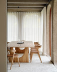 A modern dining room featuring the Ethnicraft Geometric Meeting Table in oak, six matching chairs, sheer white curtains, arched windows, and textured concrete walls creates a minimalist and airy atmosphere.