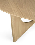 A close-up of Ethnicraft’s Geometric Dining Table – Oak, showcasing its round solid oak top and angular sculptural base with a curved cutout, all in light-colored wood.