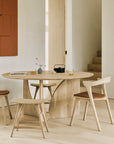 Minimalist dining area with an Ethnicraft Geometric Dining Table in oak, three wooden chairs with brown cushions, tea set, notebooks, and modern decor in a light room with beige walls and wood flooring.