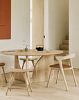 Minimalist dining area with the Ethnicraft PI Console in oak, three light wood chairs with brown cushions, a small bench, abstract red wall art, and candles for a calm, modern look.