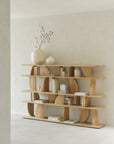 The Ethnicraft Geometric Rack - Oak, crafted in solid oak with curved, open compartments, displays books and décor against a light wall in a bright, minimalist room.