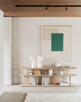 A minimalist room with an Ethnicraft Geometric Console - Oak displaying books and abstract sculptures, a white rug, modern green and beige artwork from the Geometric collection, and a wood slat ceiling above.