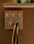 A tan coat hangs on the Ethnicraft Utilitile Wall Hanger with four vertical hooks, beneath a light wood shelf with decor, set against a brown wall—showcasing the timeless elegance of Ethnicraft design.
