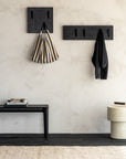 A minimalist entryway features two black Ethnicraft Utilitile Wall Hangers holding a striped tote and a black coat, complemented by a black bench with books, a round beige stool, and a neutral rug against a textured light wall.