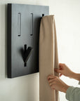A person hangs a beige fabric on the Ethnicraft Utilitile Wall Hanger, a minimalistic coat rack with geometric slots, mounted on a black rack against a white background.