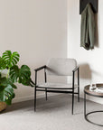 A modern room with a light gray chair, Ethnicraft Tray Coffee Table - Set of 2, a water glass, a green monstera plant in a basket, and a gray jacket on a wall hook against a white wall.