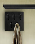 A beige coat hangs from one of four black hooks on the Ethnicraft Utilitile Wall Hanger, crafted from solid oak and mounted against a light wall with a dark horizontal shelf above.