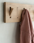 The Ethnicraft Utilitile Wall Hanger is a minimalistic, solid oak wall-mounted coat rack with angled pegs holding a pinkish-brown garment, installed on a light-colored wall.