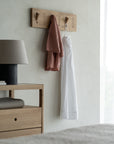 The Ethnicraft Utilitile Wall Hanger with pegs displays a rust jacket and white pants. Next to it, an Ethnicraft nightstand with a lamp and folded blanket enhances the minimal style of this bright bedroom.