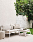 A modern patio features the Ethnicraft Donut Outdoor Pouf in Natural, a beige cushioned sofa, matching ottoman topped with a book and glass of water, all set on a light floor with lush greenery and a white brick wall as backdrop.
