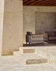 A modern patio with beige stone walls and floors features a stylish gray sofa and a partial view of the Ethnicraft Corbey Outdoor Lounge Chair under a wooden roof, with sunlight casting soft shadows across this inviting space.