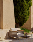 The Corbey Outdoor Lounge Chair by Ethnicraft, featuring light cushions and rope accents, sits next to a matching table with a glass bottle, creating an inviting nook beside a textured beige wall and tall cypress tree.