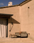 A minimalist outdoor seating area features a small sofa, Ethnicraft’s Corbey Outdoor Coffee Table, and cushions on solid teak steps by a beige stucco wall under a black awning, all bathed in warm sunlight.