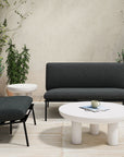 A modern patio features Moes Salma Outdoor Lounge Chairs with black quick-dry foam cushions, a matching loveseat, two round white tables, a beige rug, and potted green plants against a light wall. A cup and book sit on the coffee table.