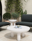 The Salma Outdoor Sofa by Moes creates a modern seating area with two dark upholstered benches, quick-dry foam cushions, white side and coffee tables topped with a cup and magazines, all surrounded by greenery and natural light.