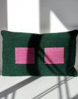 Pillow 02: Green + Purple by Ania Brzeski, a rectangular dark green pillow with two purple squares, made from locally sourced wool, sits upright on a white surface with sunlight and shadows cast on the wall and pillow.
