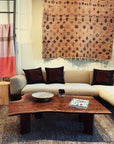 A cozy living room features a beige sofa, black and brown pillows, a patterned rug, a decorative woven wall textile, and the SOBU Bolinas Coffee Table in Claro Walnut displaying a bowl, sculpture, and magazine for modern style.