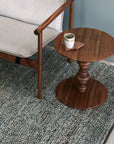 A small round wooden side table with a cup on a coaster sits next to a wooden-framed armchair with light cushions, all atop the Acacia Rug - Galapagos by Armadillo&Co, set against a light blue wall.