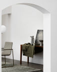 A minimal, modern interior features a woven chair, the Armadillo&Co Acacia Rug - Galapagos, and a wooden table with ceramic vases, cup, bowl, and green cloth—all set against white walls and an arched doorway.