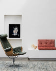 A modern minimalist living room with a green lounge chair, low white console, red-cushioned bench, table lamp, decor items, and a portrait painting in a wall niche on white walls featuring the Acacia Rug - Galapagos by Armadillo&Co.