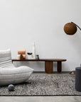 Minimalist living room featuring the Armadillo&Co Acacia Rug - Sable, a modern white lounge chair, curved floor lamp with brown shade, dark round side table, and a wooden bench with décor against a white wall.