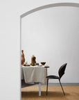 Minimalist dining setup with a black chair, white-clothed table, pottery, and wooden objects atop the Bramble Rug by Armadillo&Co. The space features a neutral palette and timeless Armadillo&Co texture framed by an arched doorway.