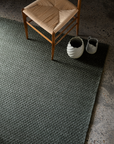 A woven chair sits on the Armadillo&Co Sherpa Rug in Kelp—a textured dark gray, hand-woven wool design—beside two decorative ceramic vases on a speckled concrete floor.