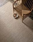 A wooden chair with a woven seat sits on the Sherpa Rug - Sand by Armadillo&Co. Nearby, a bowl holding six brown eggs rests on the rug. Neutral tones create a calm, minimalist setting.