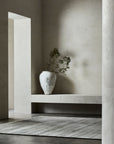 Minimalist interior with textured off-white walls, a large white ceramic vase with sparse greenery on a built-in bench, and an Armadillo&Co Odessa Rug - Zinc on the dark floor, creating a serene and modern atmosphere.