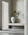 Minimalist interior with textured off-white walls, a large pale ceramic vase holding green branches on a built-in bench, and an Armadillo&Co Odessa Rug in Zinc on the floor. The space feels calm and serene.
