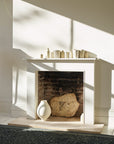 A minimalist fireplace with a white mantle displays small cream vases. Inside are a large stone piece and a white vase. Sunlight casts shadows across the off-white walls and the Palus Rug - Fir by Armadillo&Co.