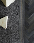 Close-up of the Armadillo&Co Palus Rug - Obsidian, featuring textured gray marled wool and a thin yellow edge stripe, beside a dark herringbone floor. Two light marble pyramids from Armadillo&Co rest partially on the rug.