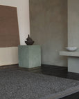 Minimalist interior with a textured Palus Rug - Obsidian by Armadillo&Co, a greenish cube pedestal holding a dark ceramic vase, neutral wall art, and a low shelf with a light bowl, all set against soft gray walls.
