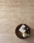 A round wooden tray with a book, black cup, and white vase with green leaves rests on the textured Staccato Rug - Latte by Armadillo&Co, most of which is shown empty in the image.