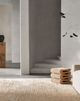 Minimalist interior with neutral tones, a curved beige sofa, sculptural wooden stool, wall art, and staircase. The space is anchored by the Armadillo&Co Staccato Rug - Latte, with a ceramic vase on a wooden pedestal near an open doorway.