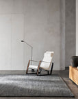 A modern minimalist room with a white rocking chair, a slim black floor lamp, and the Armadillo&Co Staccato Rug in Nightfall, plus a wooden console with ceramic vases against a gray concrete wall.